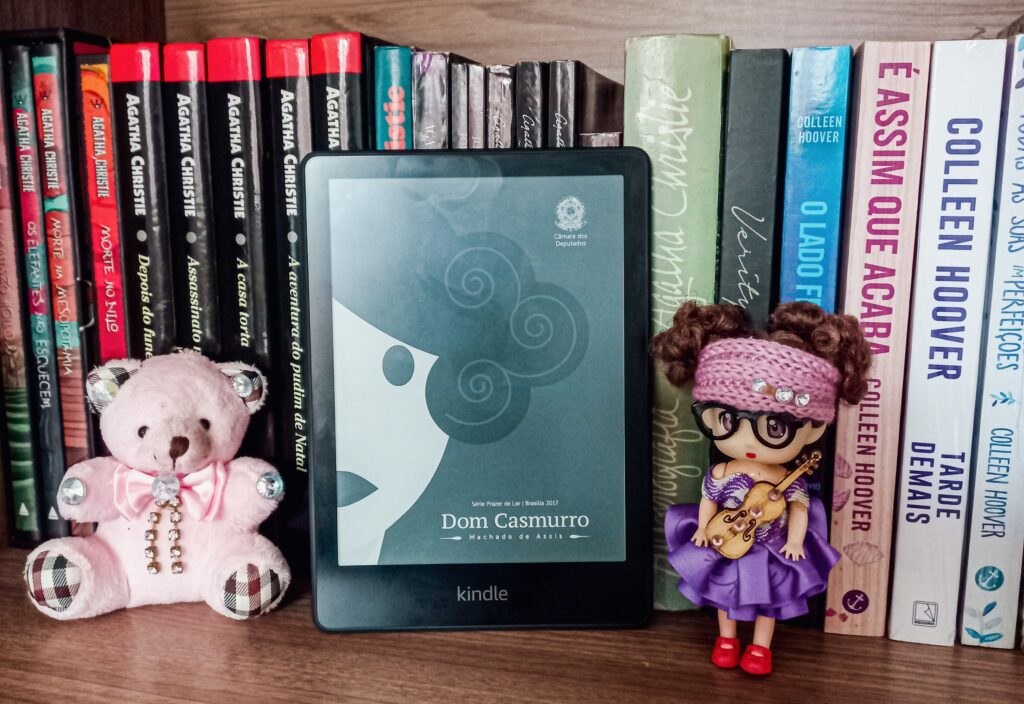 Kindle Paperwhite com a capa de Dom Casmurro em uma estante de livros e um ursinho e uma bonequinha decorativos.
